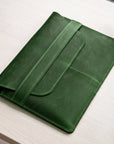 A dark green leather tablet sleeve or folio with a strap closure, shown resting on a light wood tabletop.