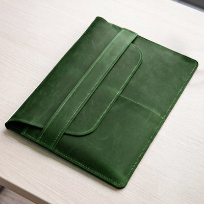 A dark green leather tablet sleeve or folio with a strap closure, shown resting on a light wood tabletop.