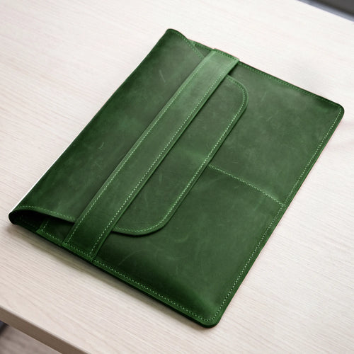 A dark green leather tablet sleeve or folio with a strap closure, shown resting on a light wood tabletop.