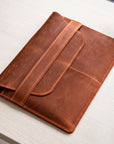 A brown leather tablet sleeve or folio with a strap closure, photographed on a light wood tabletop.