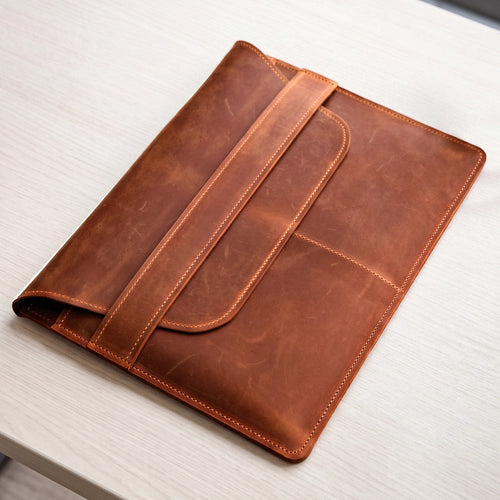 A brown leather tablet sleeve or folio with a strap closure, photographed on a light wood tabletop.