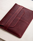 A dark red leather sleeve or folio with a strap closure, photographed on a light wood tabletop.