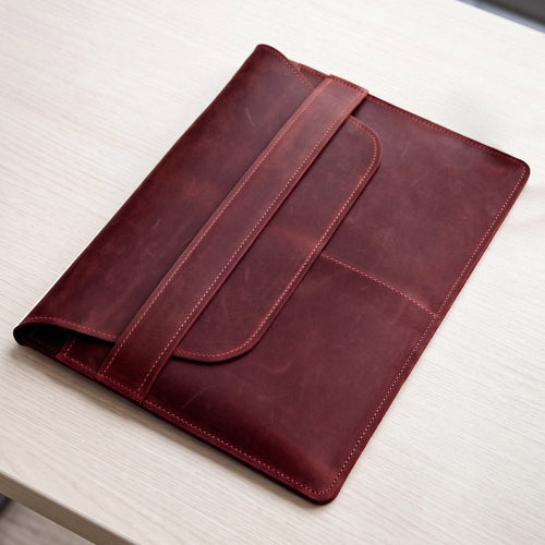 A dark red leather sleeve or folio with a strap closure, photographed on a light wood tabletop.