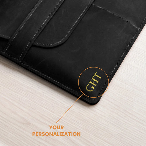 Close-up of a black leather folio or laptop sleeve on a light wood surface, with an orange callout pointing to the corner where the letters 'GHT' are personalized, and text reading 'YOUR PERSONALIZATION'.