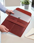 A person in a light blue dress shirt holds a brown leather laptop sleeve containing a silver laptop on a light-colored desk with a pen and papers nearby.