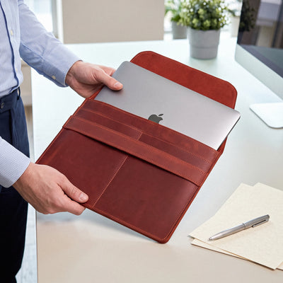A person in a light blue dress shirt holds a brown leather laptop sleeve containing a silver laptop on a light-colored desk with a pen and papers nearby.