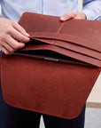 A person holds open a brown leather tablet sleeve or document cover, showing card slots and a tablet inside. The person is wearing a light blue striped shirt and dark pants in a bright indoor setting.