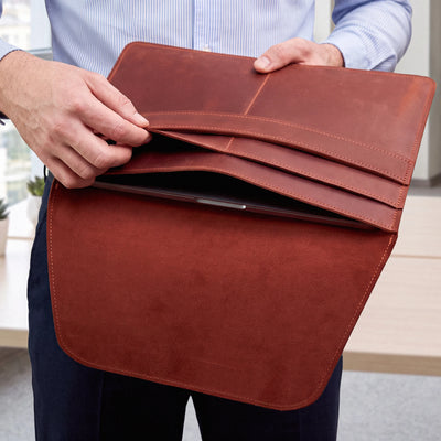 A person holds open a brown leather tablet sleeve or document cover, showing card slots and a tablet inside. The person is wearing a light blue striped shirt and dark pants in a bright indoor setting.