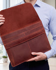 A person in a light blue striped shirt holds up a large brown leather portfolio folder or document sleeve in an indoor office setting.