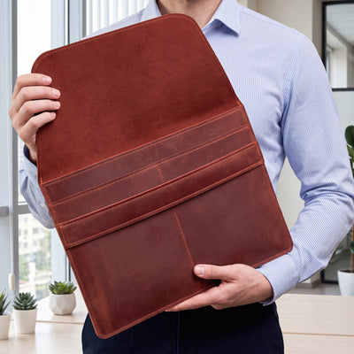 A person in a light blue striped shirt holds up a large brown leather portfolio folder or document sleeve in an indoor office setting.