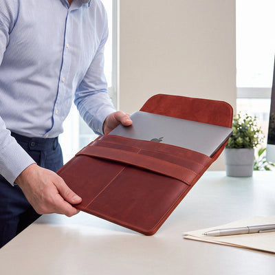 A person in a light blue dress shirt holds an open reddish-brown leather laptop sleeve with a closed silver laptop inside, set in a bright office environment with a desk, notebook, and plant in the background.
