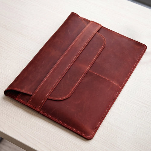 A reddish-brown leather folio or tablet sleeve with a strap closure, photographed on a light wood tabletop.