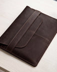 A dark brown leather folio or tablet sleeve with a strap closure, shown resting on a light wood tabletop.