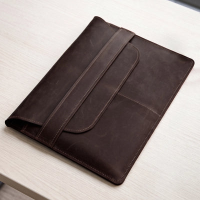 A dark brown leather folio or tablet sleeve with a strap closure, shown resting on a light wood tabletop.