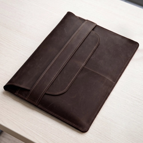 A dark brown leather folio or tablet sleeve with a strap closure, shown resting on a light wood tabletop.