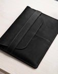 A black leather tablet sleeve or folio case with a strap closure, resting on a light wood tabletop.