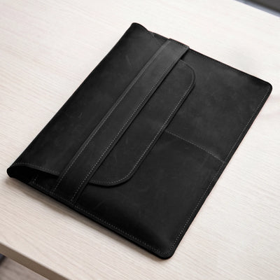 A black leather tablet sleeve or folio case with a strap closure, resting on a light wood tabletop.
