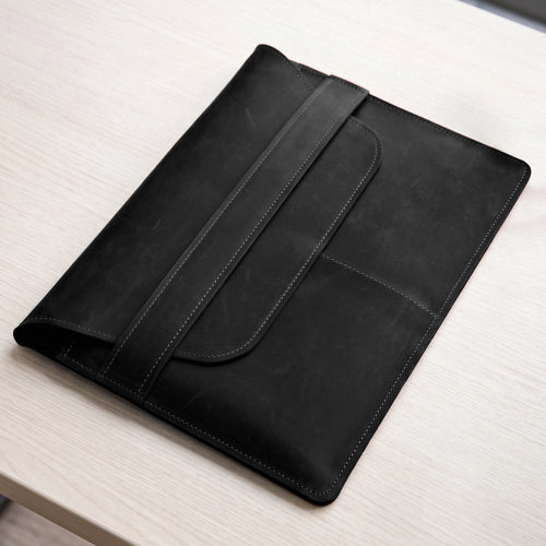 A black leather tablet sleeve or folio case with a strap closure, resting on a light wood tabletop.
