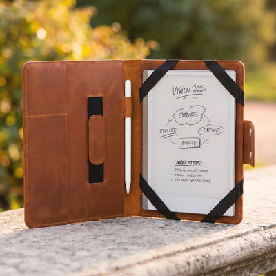 A brown leather tablet case stands open on a stone surface outdoors, holding a white tablet with black elastic bands and a white stylus inside. The background is a blurred natural outdoor setting with green and yellow foliage.