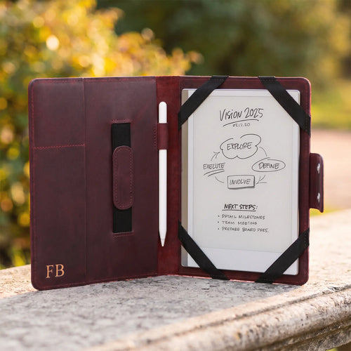 A burgundy leather tablet case shown open on a stone surface outdoors, with a white tablet inserted and black elastic bands holding it in place. The case has card slots on the left, a pocket tab, and a gold 'FB' monogram near the bottom left.