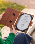 A person in a green sweater holds an open brown leather tablet case outdoors. The case has black elastic bands and a white tablet inside with handwritten notes visible.