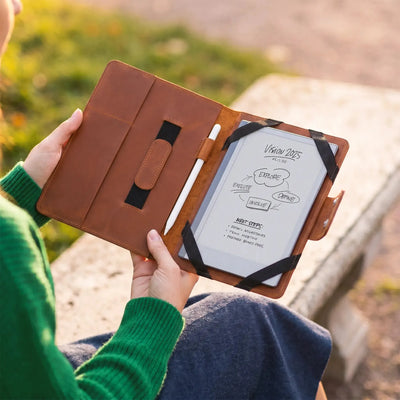 A person in a green sweater holds an open brown leather tablet case outdoors. The case has black elastic bands and a white tablet inside with handwritten notes visible.