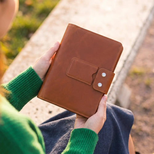 A person in a green sweater holds a brown leather journal or notebook with a snap-button strap closure, photographed outdoors on a sunny day.