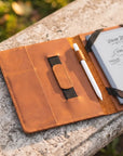 A tan leather notebook cover or organizer laid open on a stone surface, holding a white stylus and a small notebook. The cover has stitched pockets, a black elastic closure, and visible sketch notes on the inside page.