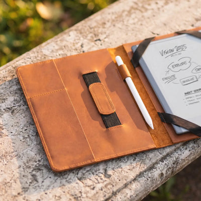 A tan leather notebook cover or organizer laid open on a stone surface, holding a white stylus and a small notebook. The cover has stitched pockets, a black elastic closure, and visible sketch notes on the inside page.
