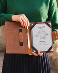 A person in a green sweater holds an open brown leather planner or notebook cover. The cover has black ribbon-like straps, a pocket with a brown tab, and a white sheet inserted with handwritten notes and a pen.