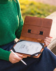 A person in a green sweater holds an open brown leather tablet sleeve on their lap outdoors, with a tablet inside and a white stylus in hand.