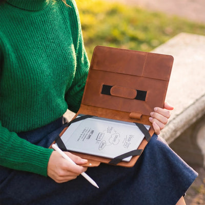 A person in a green sweater holds an open brown leather tablet sleeve on their lap outdoors, with a tablet inside and a white stylus in hand.