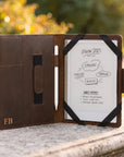 A brown leather tablet case stands open on a stone surface outdoors, with a white tablet inserted and black elastic straps holding the cover open. The case has a small gold 'FB' logo on the lower left and a vertical magnetic closure tab on the left interior.