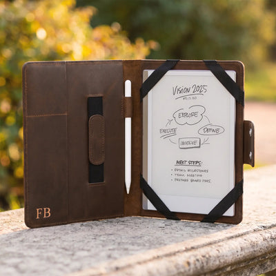 A brown leather tablet case stands open on a stone surface outdoors, with a white tablet inserted and black elastic straps holding the cover open. The case has a small gold 'FB' logo on the lower left and a vertical magnetic closure tab on the left interior.