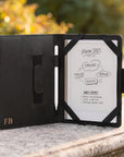 A black tablet case with a white screen insert and stylus, photographed open on a stone surface outdoors with blurred greenery in the background. The case has a small gold 'FB' logo on the lower left.