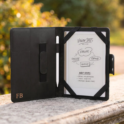 A black tablet case with a white screen insert and stylus, photographed open on a stone surface outdoors with blurred greenery in the background. The case has a small gold 'FB' logo on the lower left.