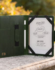 A black tablet case with a white screen insert and stylus, photographed open on a stone surface outdoors with blurred greenery in the background. The case has a small gold 'FB' logo on the lower left.