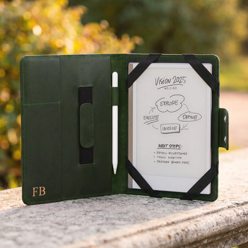 A black tablet case with a white screen insert and stylus, photographed open on a stone surface outdoors with blurred greenery in the background. The case has a small gold 'FB' logo on the lower left.