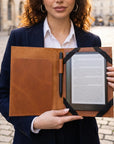 A woman in a navy blazer and white shirt holds an open brown leather e-reader case with a pen and a visible Kindle e-reader inside, set against a blurred cobblestone street background.