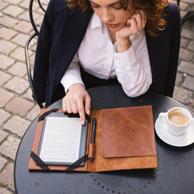 A woman in a navy blazer and white shirt sits at an outdoor table, looking down at an open brown leather folio case containing an e-reader, a pen, and a closed matching leather cover. A cup of coffee is on the table beside her.
