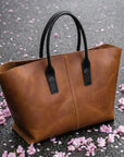 Brown leather tote bag with black handles on a gray surface with pink flower petals.