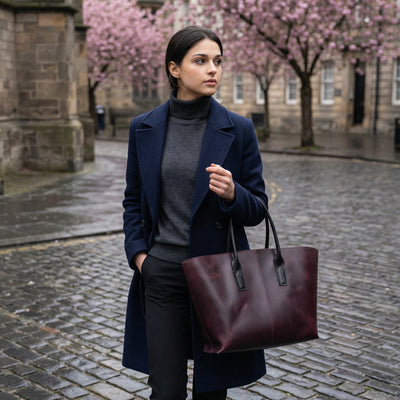 Woman in a blue coat holding a maroon bag on a cobbled street with cherry blossom trees in the background.