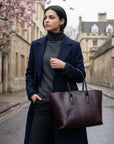 Woman holding a brown leather tote bag on a street with cherry blossoms and stone buildings.