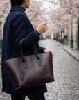 Person holding a brown leather tote bag on a street with cherry blossom trees.